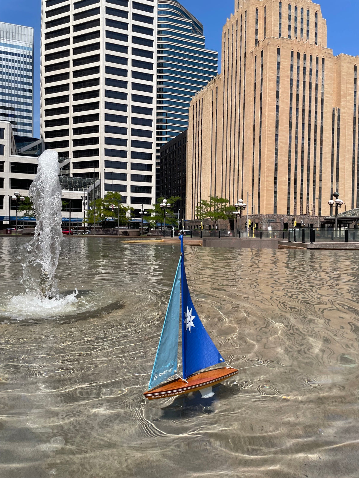 A small wooden toy sailboat with blue and teal sails floats on the shallow water of an urban fountain plaza, with modern skyscrapers in the background.