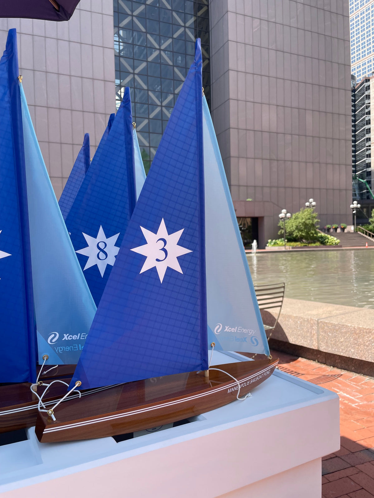 Several blue and white model sailboats with large star-shaped markings on the sails are displayed on a white pedestal in an outdoor urban setting with a modern glass building and water feature in the background.