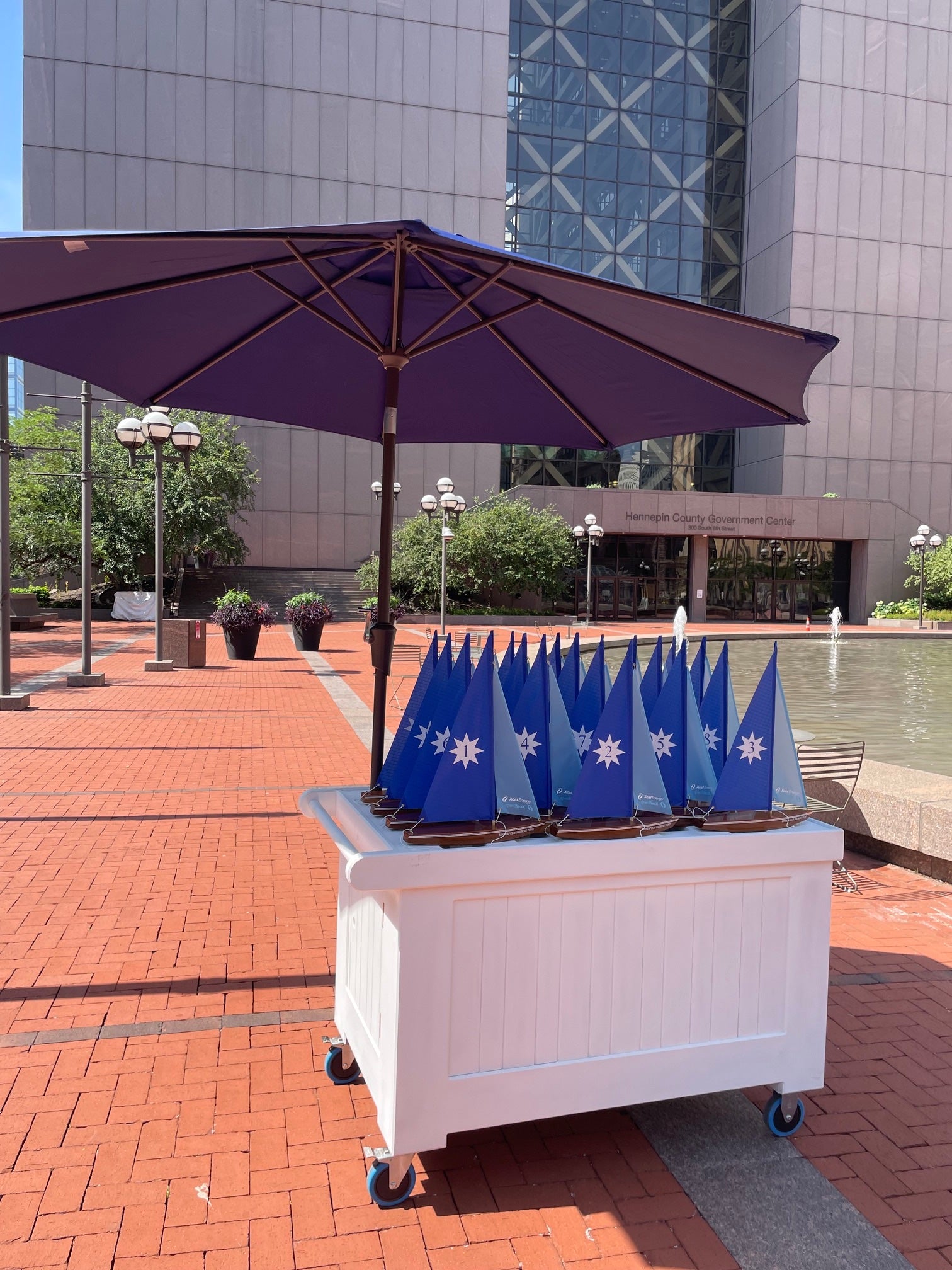 A white rolling display cart holding multiple blue flags with white snowflake-like logos, set under a large purple patio umbrella on a red brick plaza outdoors.