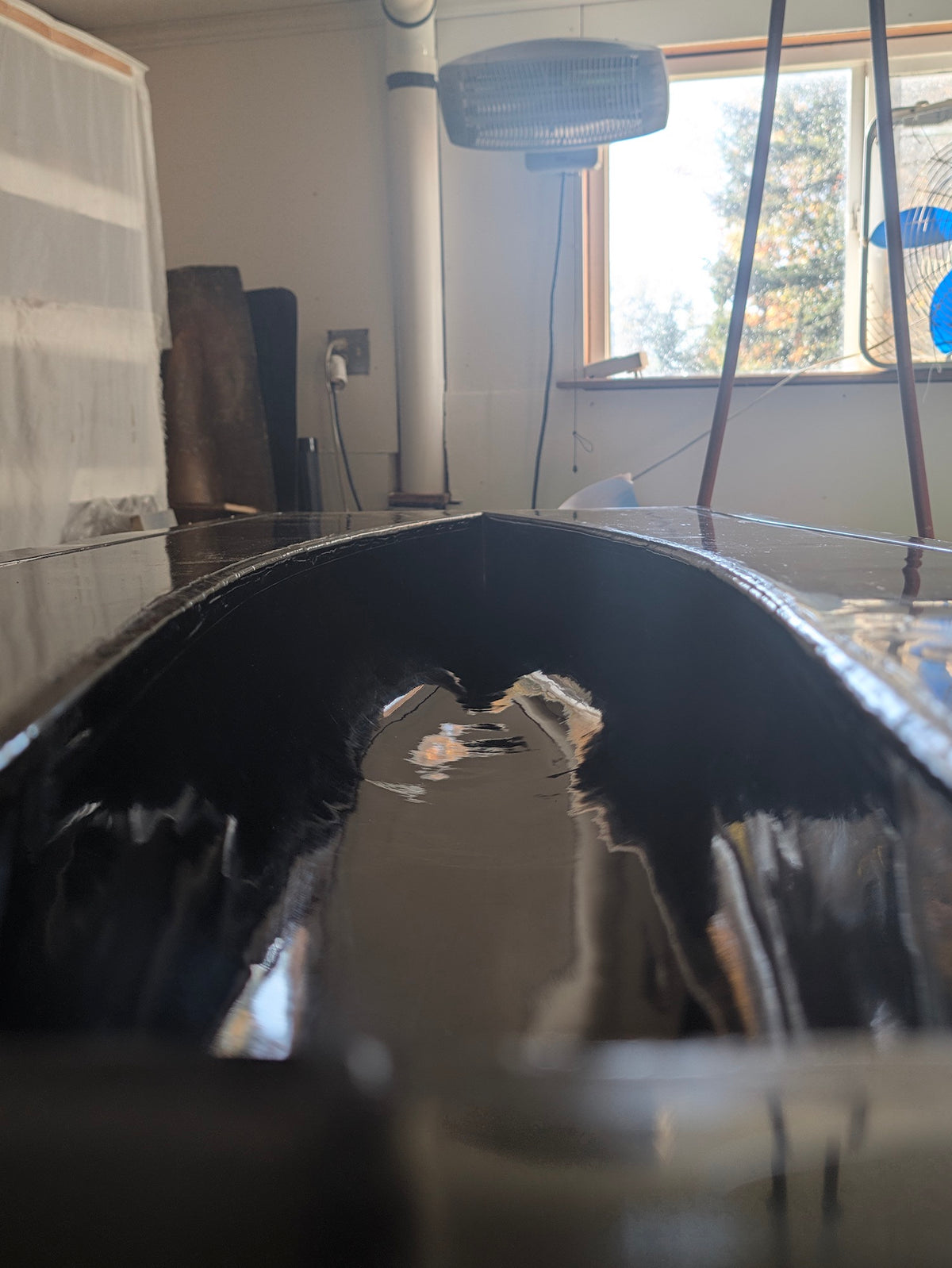 A close-up view inside a glossy black boat or hull, showing reflections of the surrounding workshop environment, including a window, fan, and floor.
