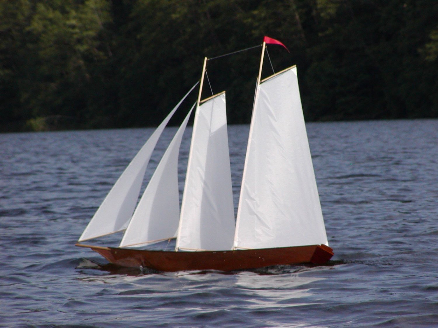 A radio-controlled mahogany plywood hull sailboat with two masts and white sails, on water.